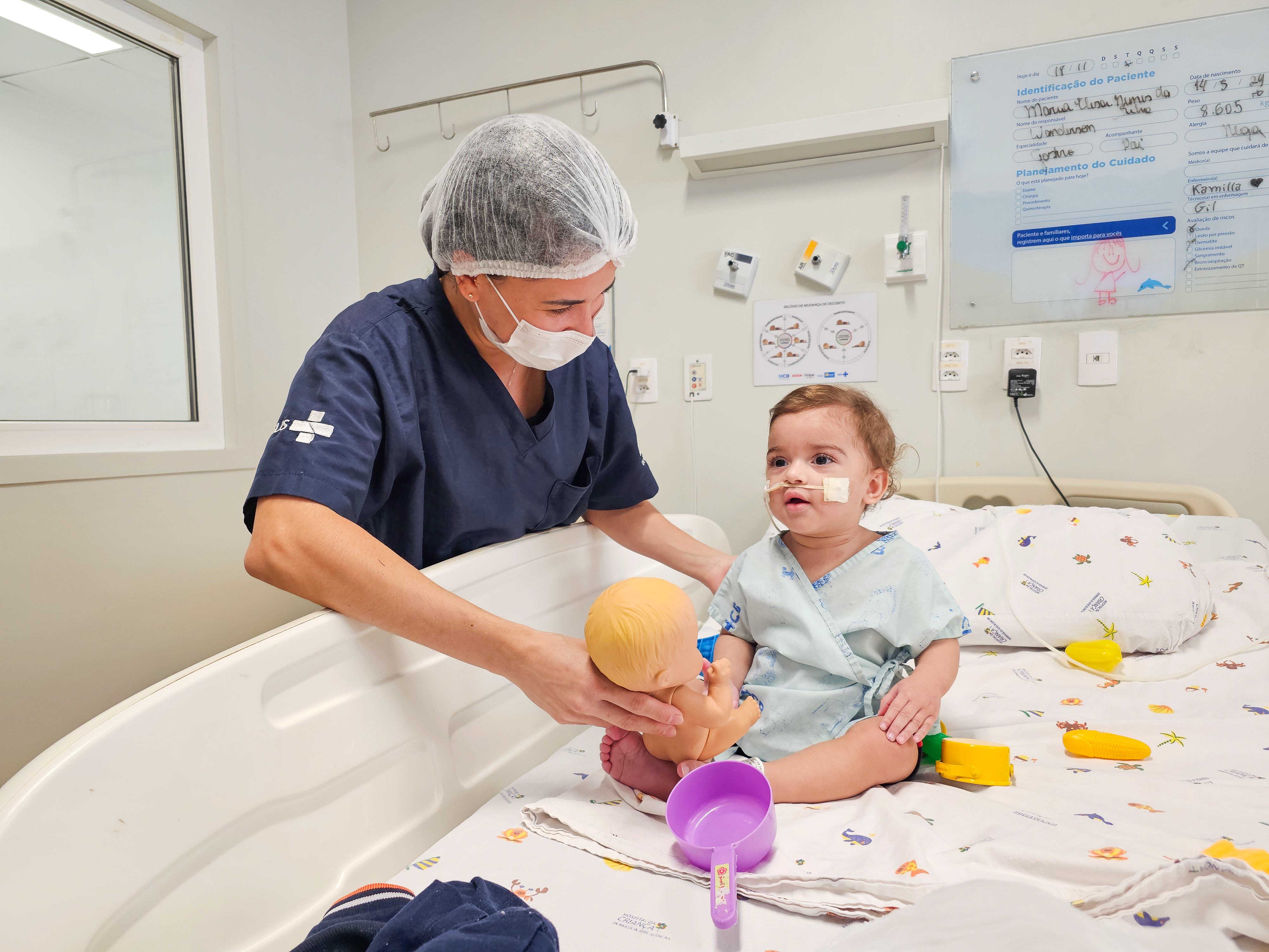 Foto de atendimento no leito. Há uma mulher de uniforme azul, touca e máscara hospitalar segurando um boneco diante de um bebê, que está sentado na cama. O bebê veste roupa hospitalar verde-claro, e está com sonda no nariz enquanto olha para frete. O quarto tem paredes claras com alguns dizeres não identificados na parte de trás.