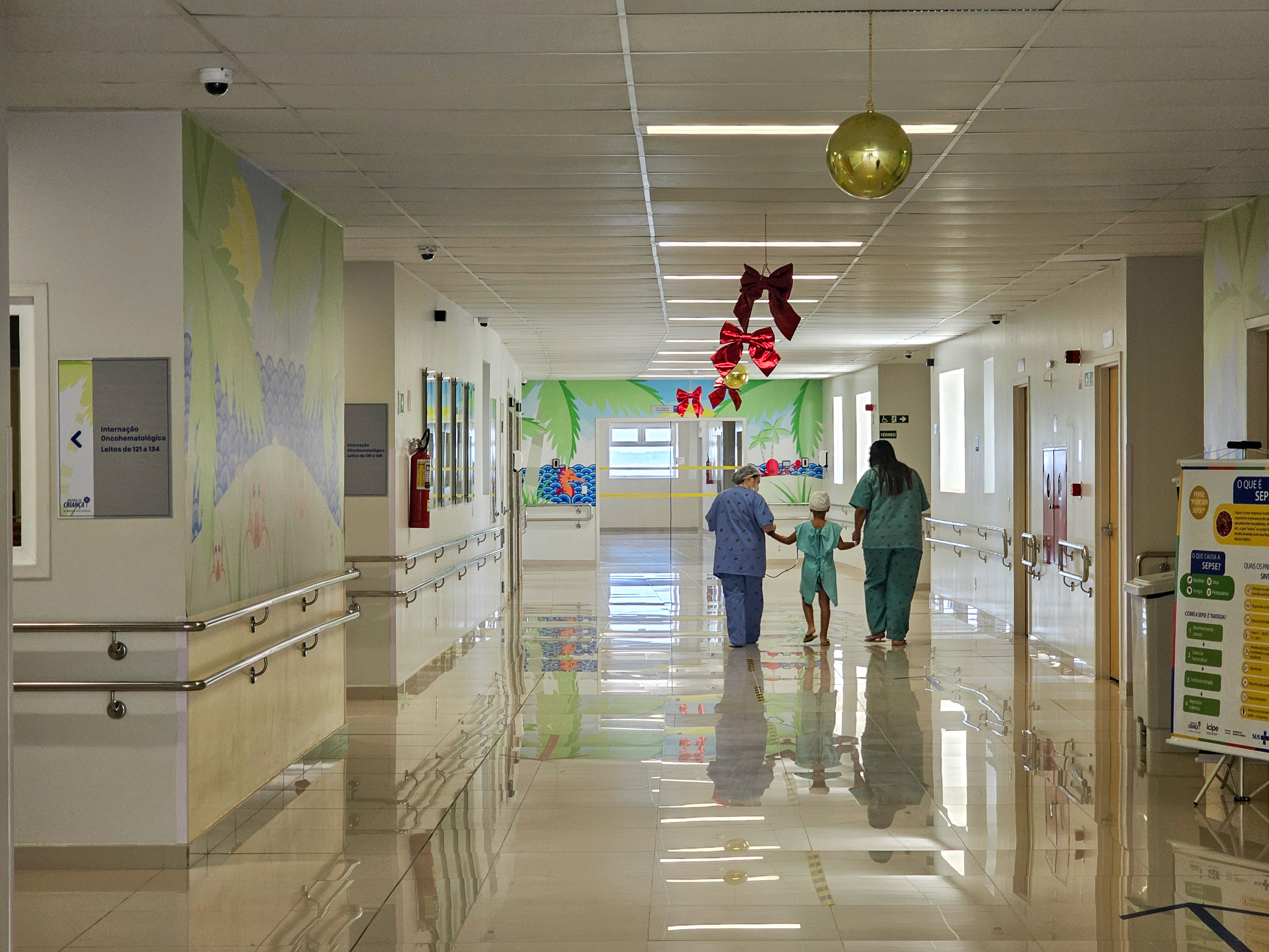 Foto em plano geral do corredor do Hospital da Criança de Brasília. Paredes e teto branco, com alguns adesivos coloridos em algumas paredes. Ao fundo, mais à direita, há três pessoas de costas andando, duas adultas e uma criança, de mãos dadas. 