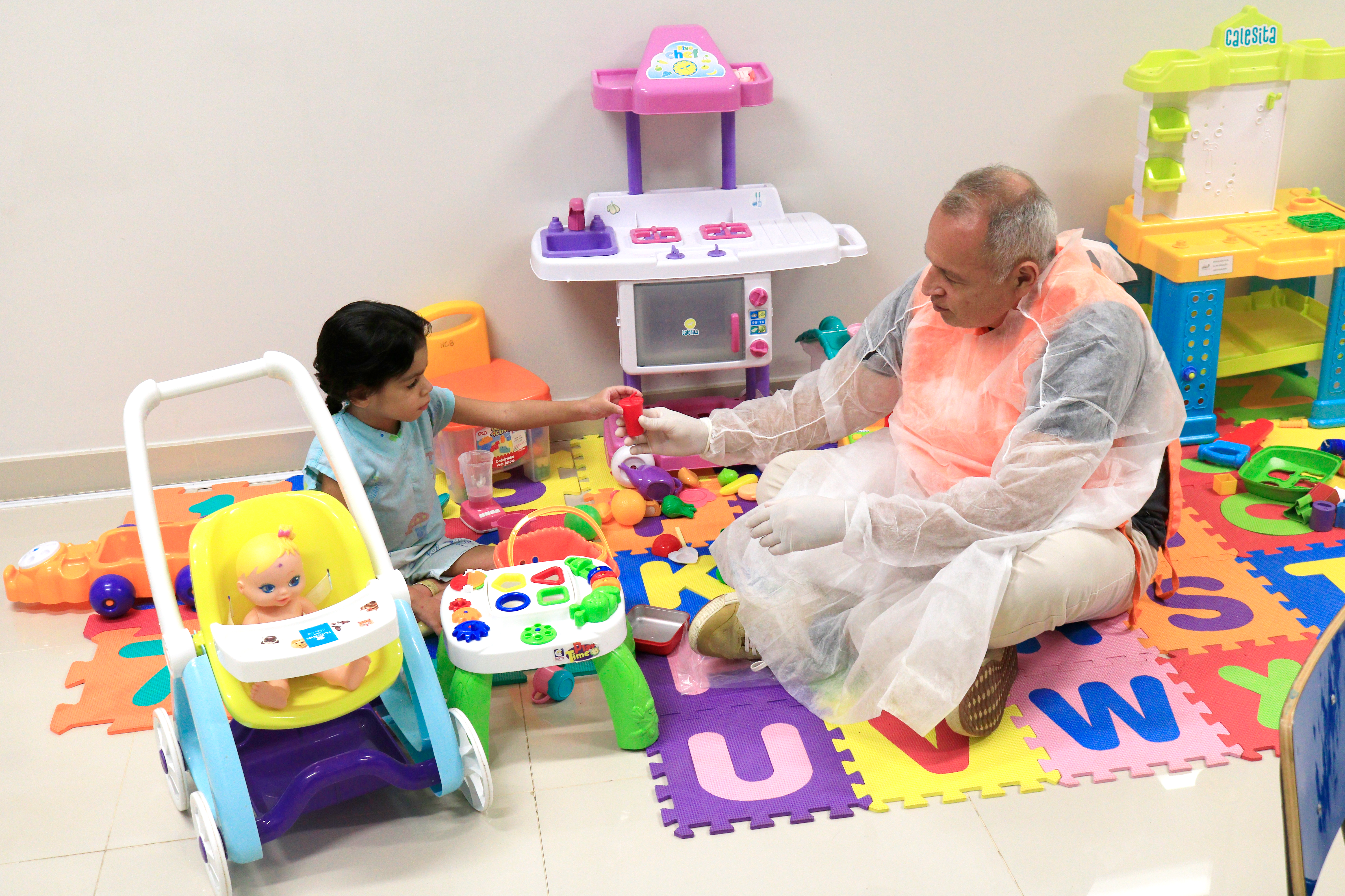 Foto em plano geral de um adulto e uma criança brincando em uma brinquedoteca. A criança é uma menina com roupa hospitalar e cabelos escuros e trançados, que está de frente a uma homem idoso com capote hospitalar (tecido transparente por cima da roupa. Eles estão sentados em cum tapete colorido e há brinquedos espalhados ao redor deles.